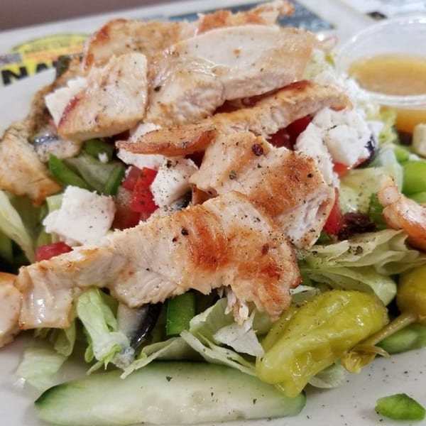 Grilled chicken salad with lettuce, tomatoes, cucumbers, feta cheese, and peppers, with dressing on the side.