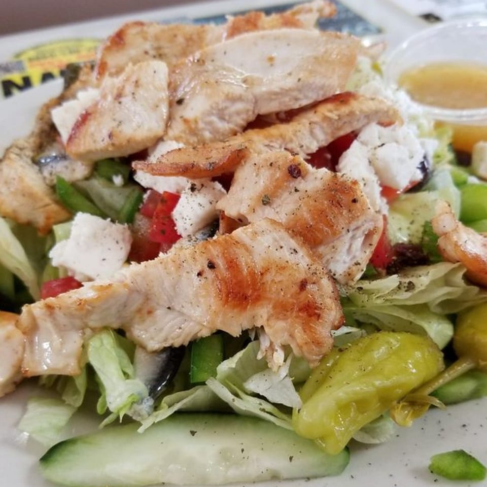 Grilled chicken salad with lettuce, tomatoes, cucumbers, feta cheese, and peppers, with dressing on the side.